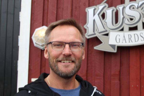 en leende man med glasögon framför en vägg där delar av en logotype för Kustens Gårdsbryggeri syns